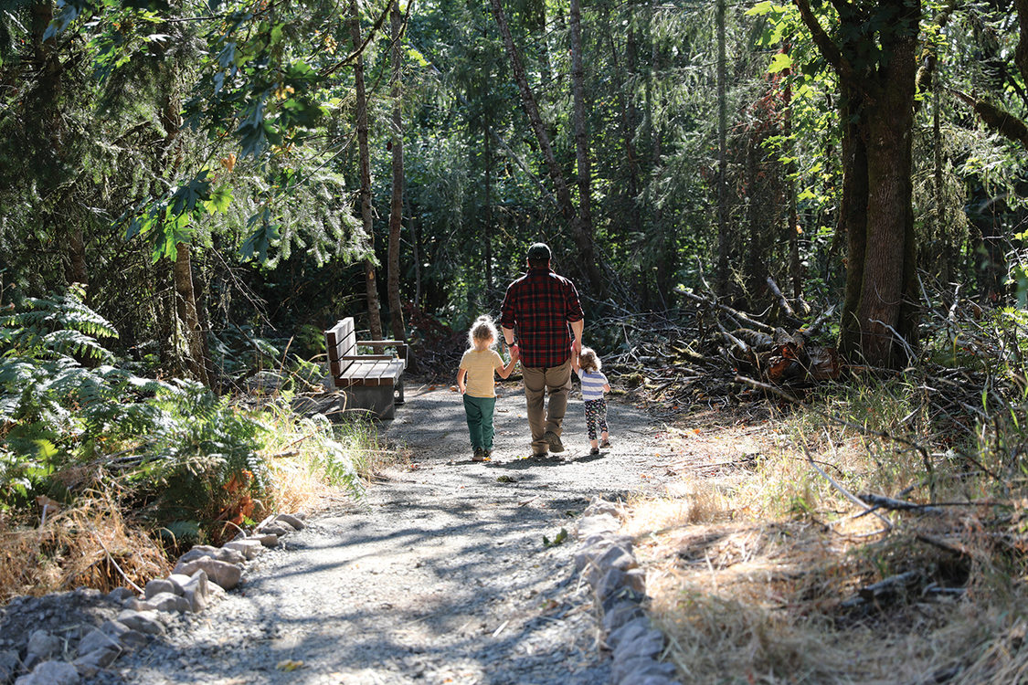An adult holds hands and walks with two little kids down a trail.