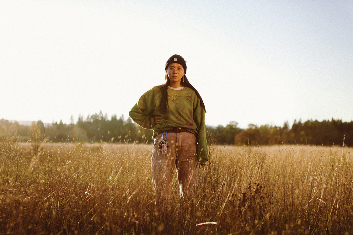 A person with long black hair stands in a field of brown grass, their hand is on their hip and they look straight at the camera.