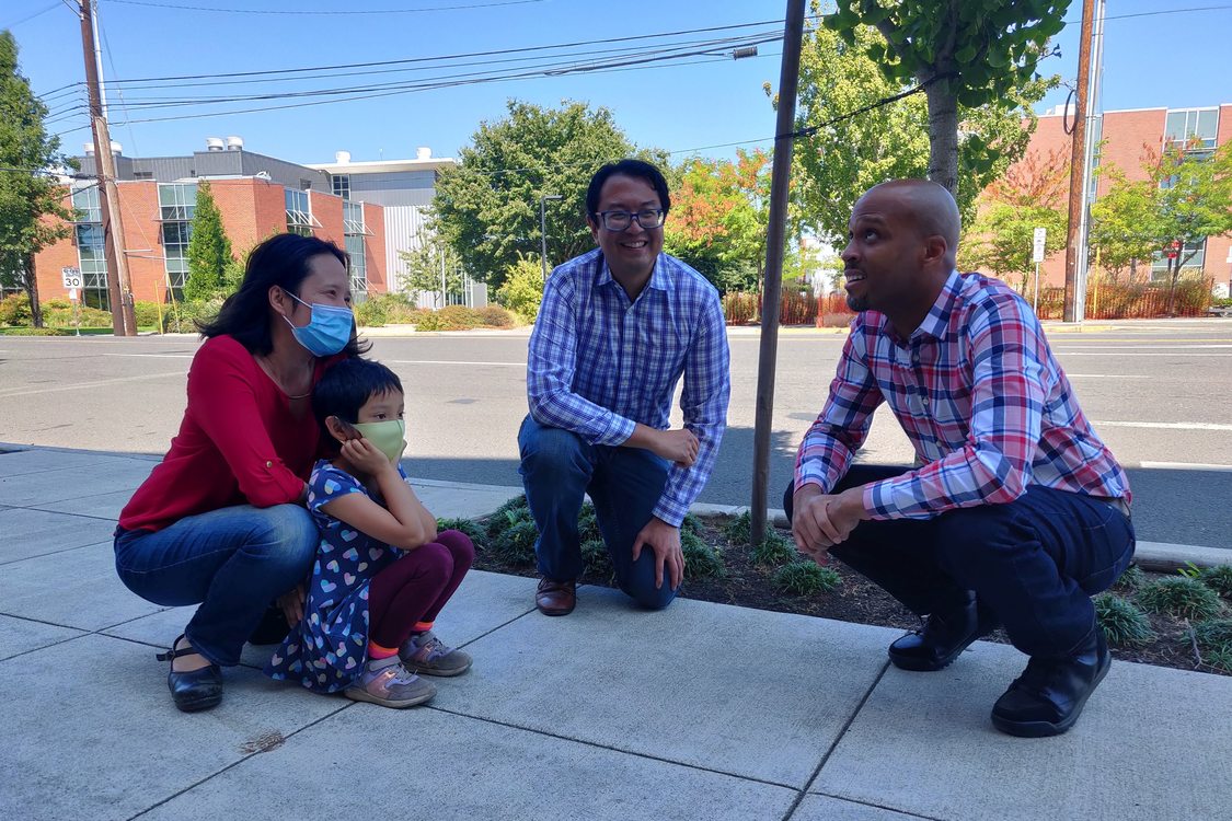 Metro Councilor Duncan Hwang crouching on knees alongside a masked woman and child and a man wearing a plaid shirt on the sidewalk next to the affordable housing development Orchards of 82nd Avenue