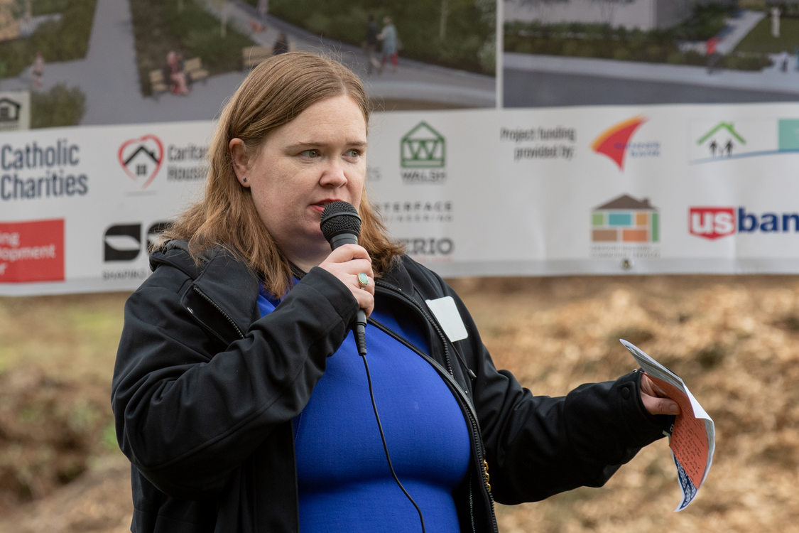 Metro Councilor Christine Lewis holding a microphone and carrying a paper pamphlet, addressing a crowd at the groundbreaking of Good Shepherd Village affordable housing project in Happy Valley, Oregon