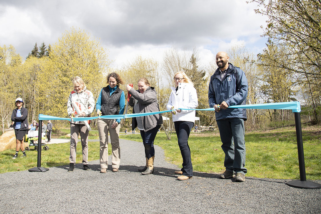 Five people stand in a row holding scissors and are ready to cut a teal ribbon. The person in the middle holds oversized scissors.