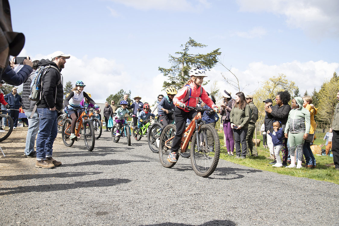 Children on mountain bikes ride on a gravel trail between two rows of onlookers.