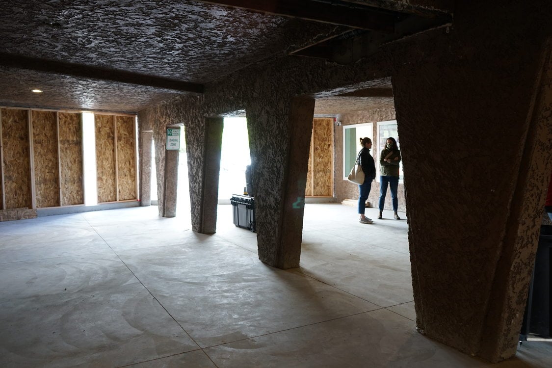 Interior photo of a common area in Aloha Inn. The walls are exposed framing and the floor is bare concrete at this stage of construction. Two people on a construction tour of the property are visible in the background.