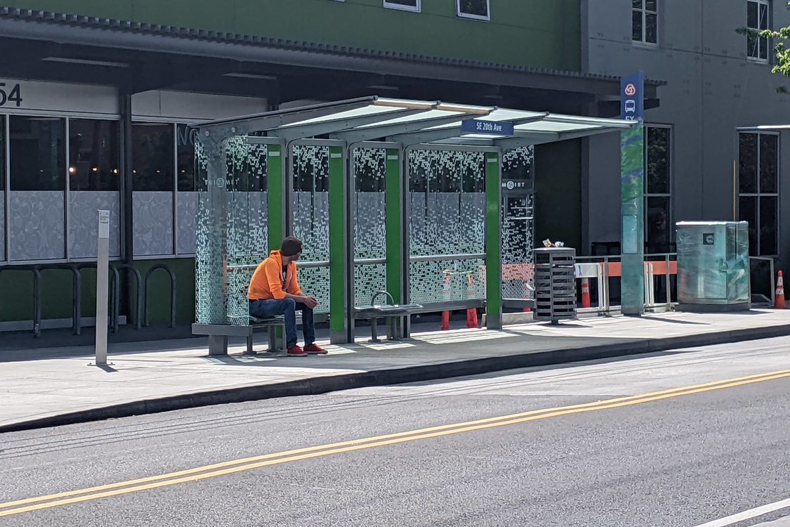 A man wearing a bright orange hoodie and red sneakers sits on a bench under a transit shelter on Southeast 20th Avenue in Portland