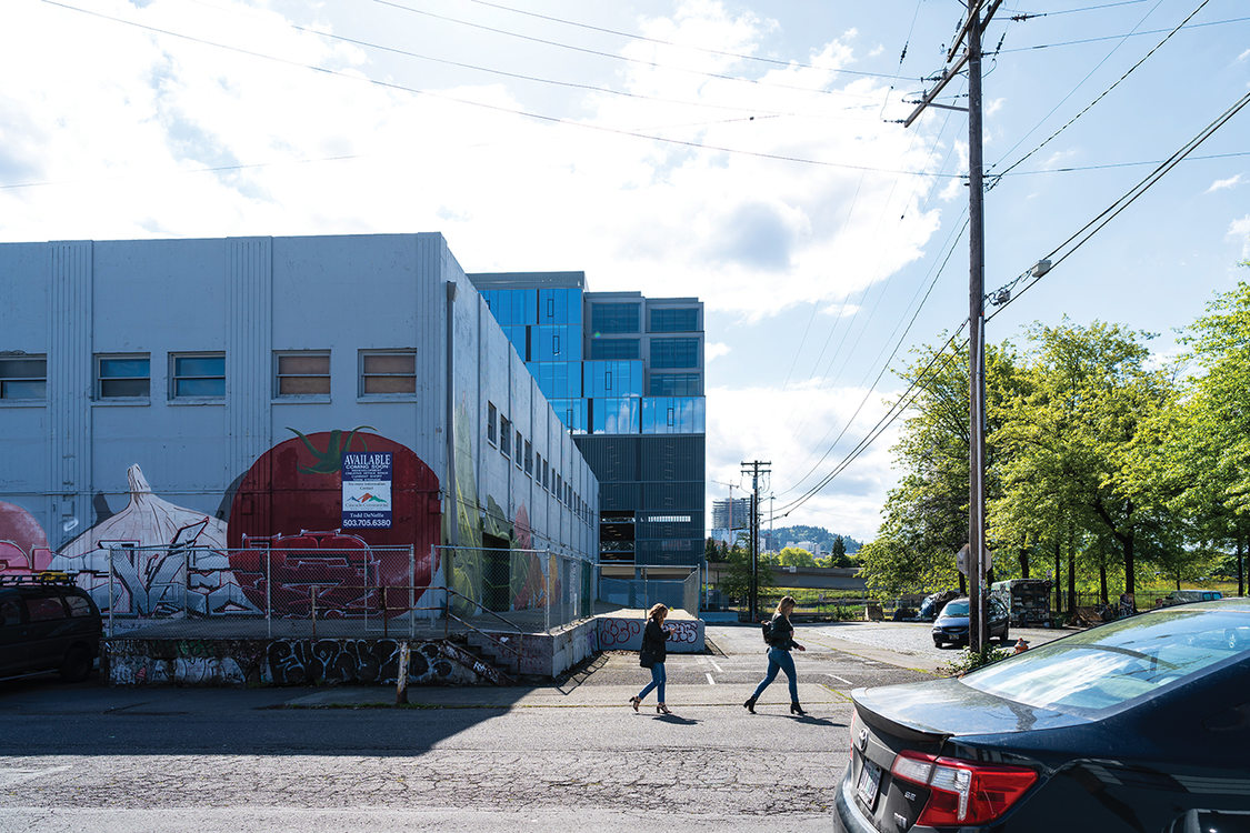 The sun glares off of the road in a downtown industrial neighborhood. Two people walk in the bright light. A few trees provide some shade down the road.