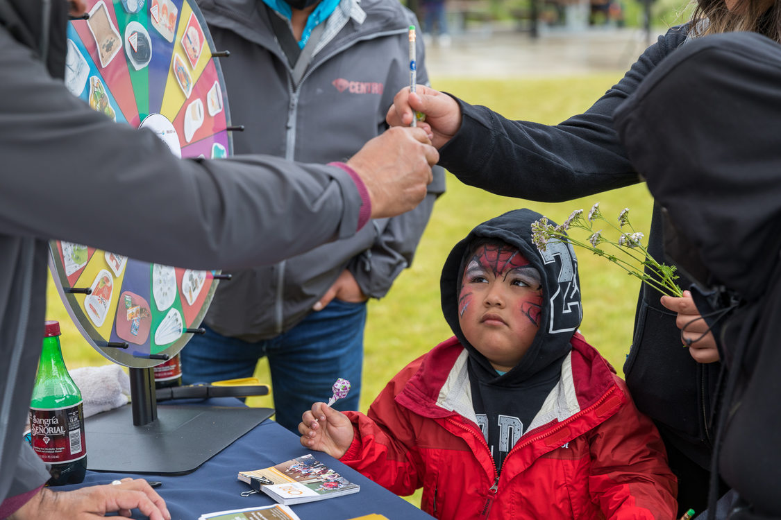 A little boy in a rain jacket, face painted like Spider-Man, holds a lollipop while adults above him talk at an informational table.