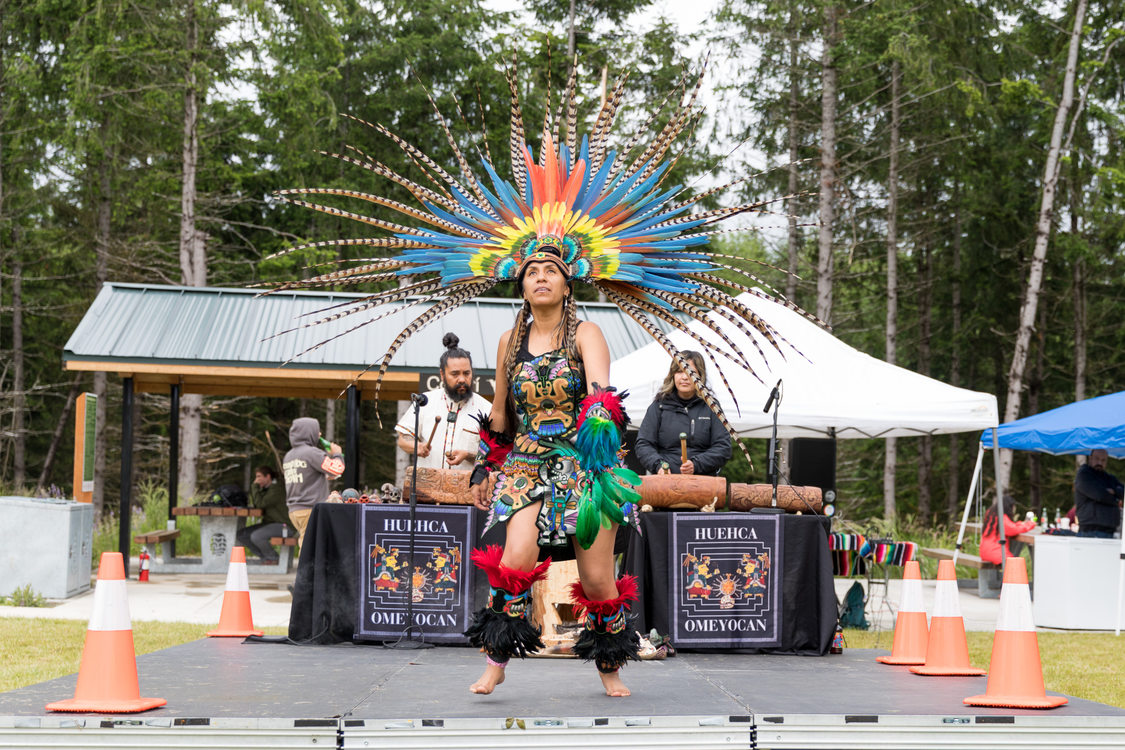 A woman wearing traditional Indigenous Mexican regalia dances on a stage at a park