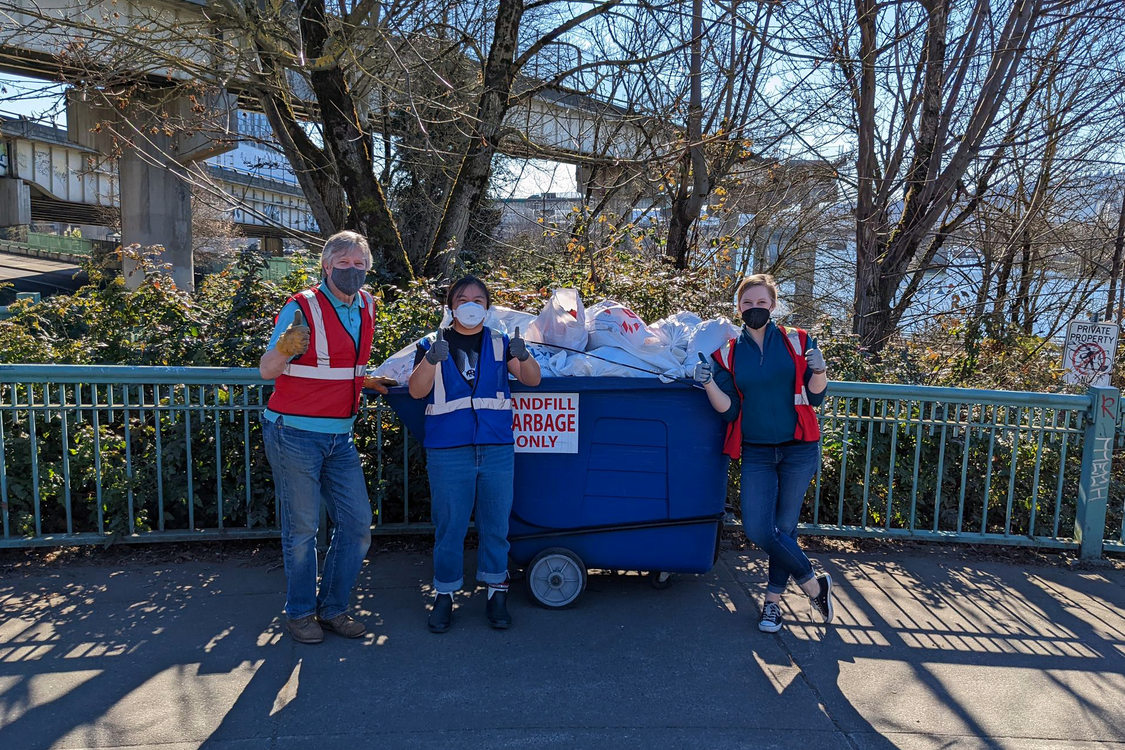 An image of three volunteers giving the thumbs up sign, dressed in safety vests in front of a small dumpster 