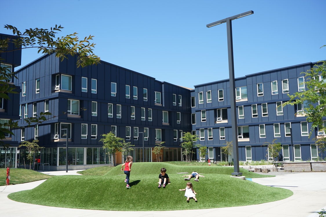 Children playing on a grass mound in front of dark blue multifamily housing