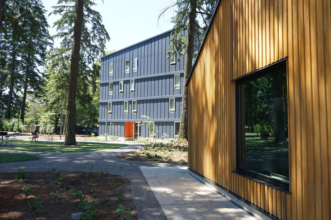 Wood paneled building in the right part of the frame, with dark blue multifamily building and tall trees in the background