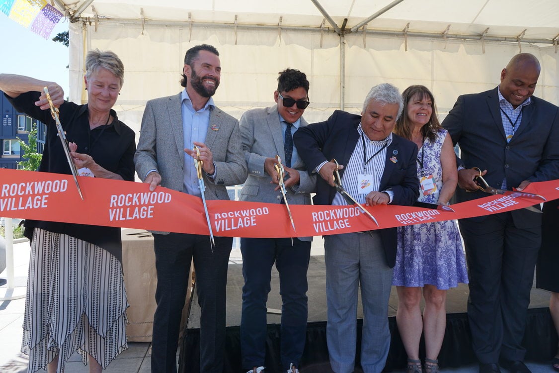 Six people standing and cutting a red ribbon that says "Rockwood Village" with oversized scissors