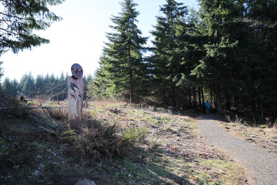 Statue on the left of trail that leads into a forested area of doug fir in the background.