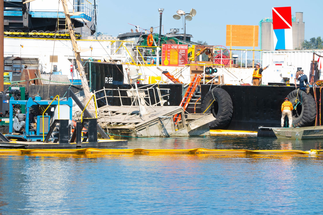A derelict sunken vessel is raised from the Columbia River by a larger Coast Guard salvage vessel. Several crew members wearing safety gear direct the salvage efforts.
