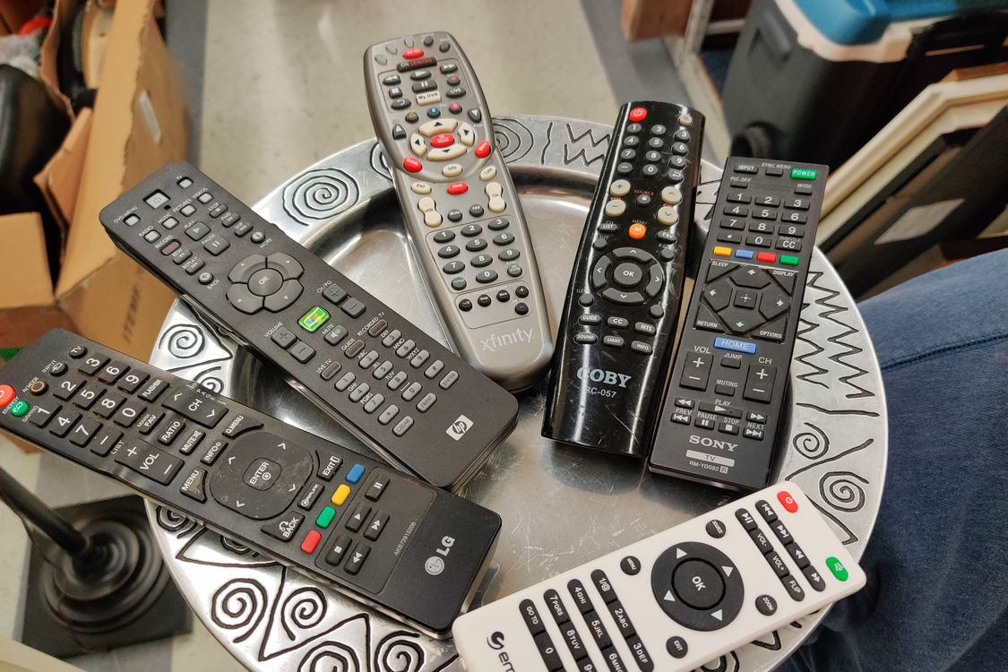 Used remotes places on a metal plate. The remotes are arranged in an array, splaying out from the center of the plate, except for one white remote near the bottom of the image.