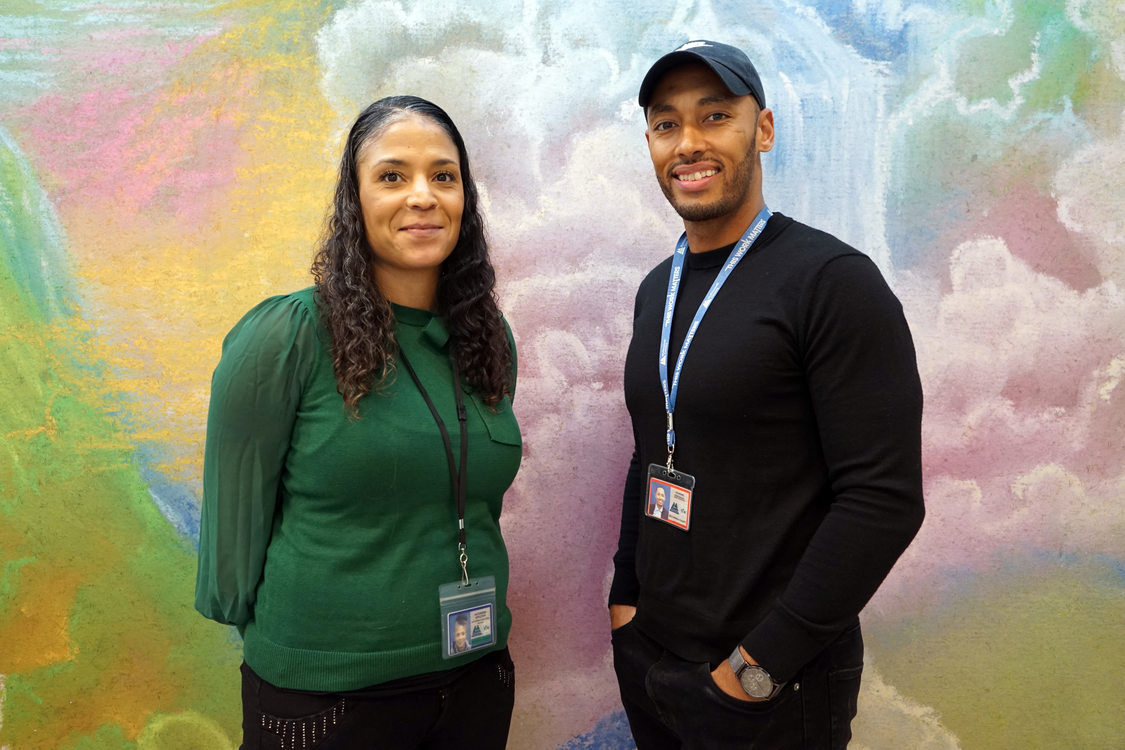 A Black man and woman standing in front of a colorful pastel mural, smiling