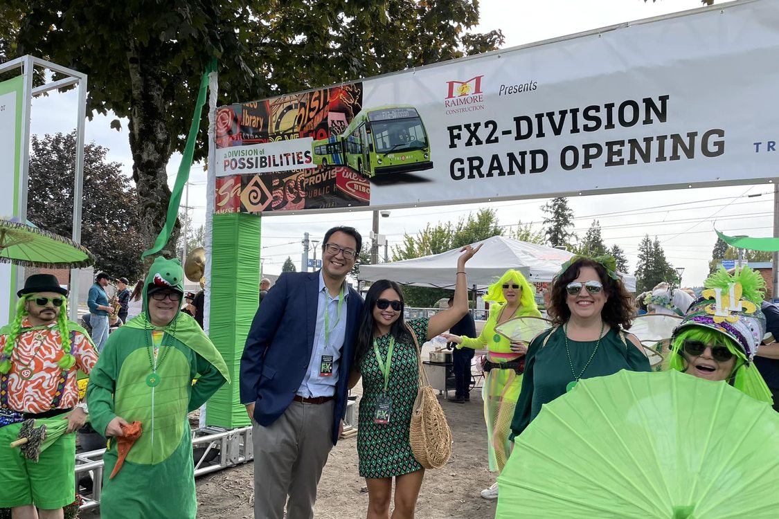 Metro Councilor Duncan Hwang stands next to Kathy Wai under a sign banner displaying "FX2-Division Grand Opening" next to many people dressed in bright green costumes of many styles.