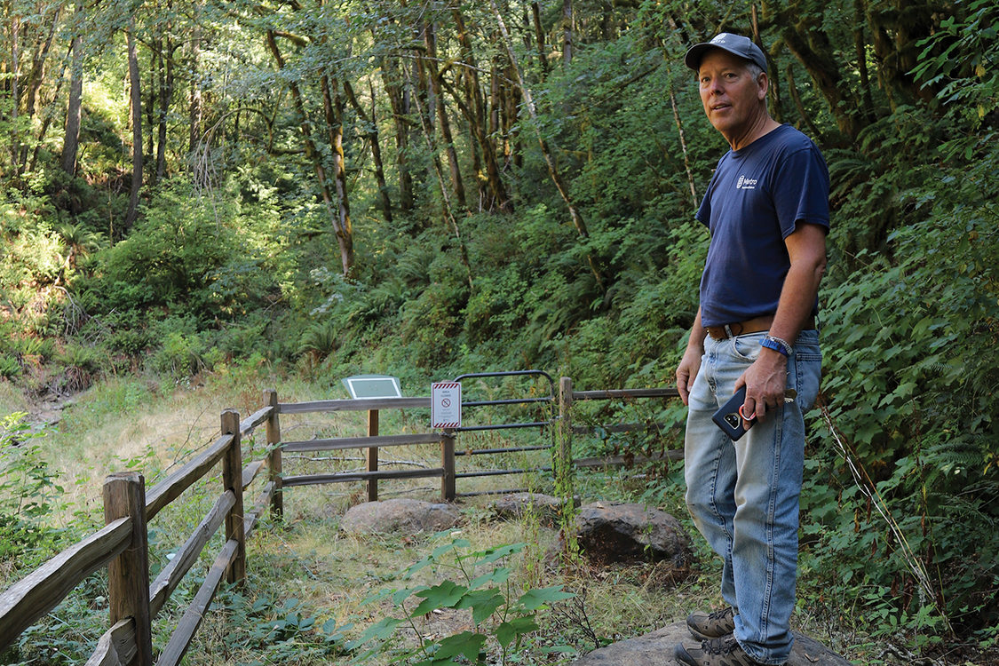 A man in a T-shirt and jeans stands on a boulder overlooking a forest clearing behind wooden fences.