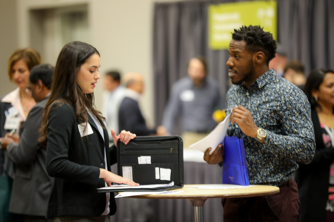 A woman with long dark brown hair, wearing a black blazer, rests her hand on papers in an open notebook while speaking to a Black man wearing a floral print shirt and carrying a swag bag at Metro's small business open house networking event.