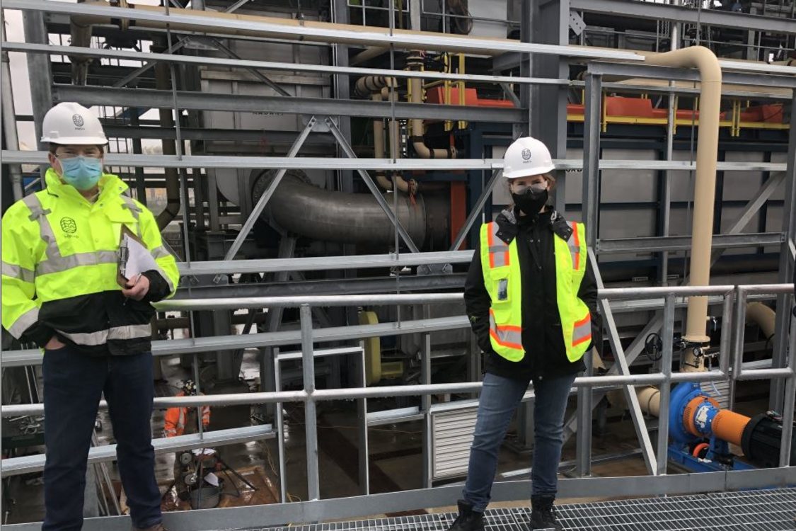 Metro solid waste enforcement inspectors Will Ennis and Hila Ritter are wearing hard hats, yellow safety jackets, and masks, and stand next to an autoclave inside a transfer station in greater Portland.