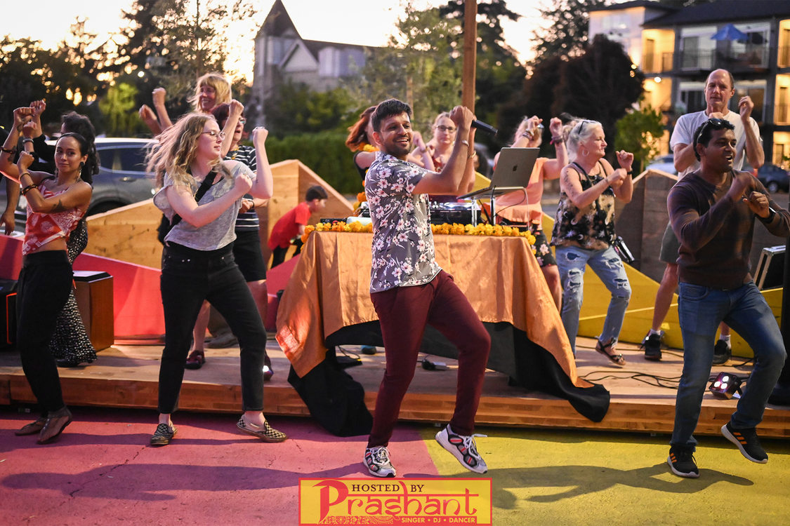 A group of people of all ages dance at sunset in front of a stage set up in the middle of a neighborhood