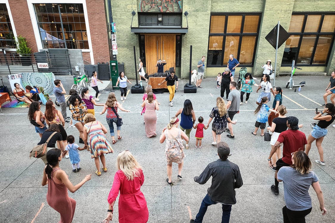 A large group of people of all ages dance in the street in front of a DJ