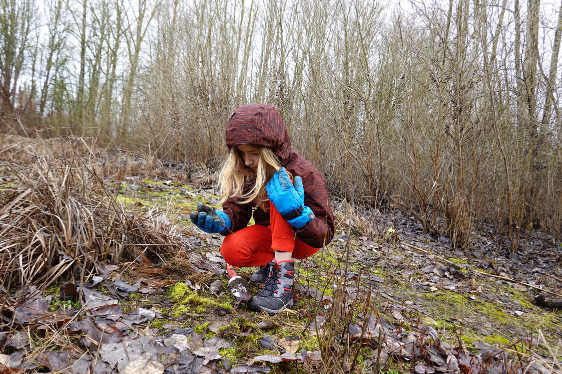 12-year-old boy with long hair in coat and gloves crouches in a wetlands area and examines a small sedge plant in his hand