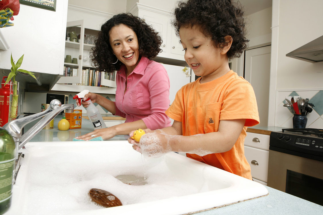 A woman and her son lean over a kitchen counter. The son plays with bubbles in the sink while the mom sprays down the countertops