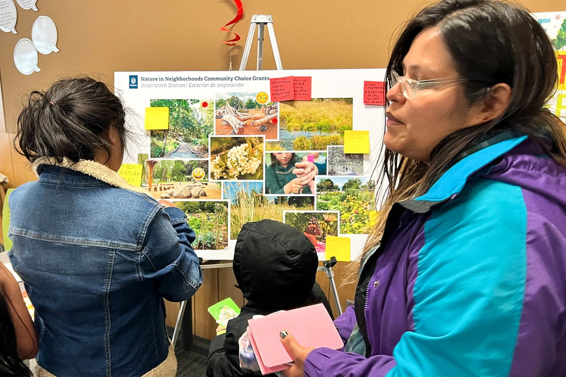 Two women and a child look at a board with different pictures of outdoor spaces. Sticky notes are attached to the board with comments from community members.