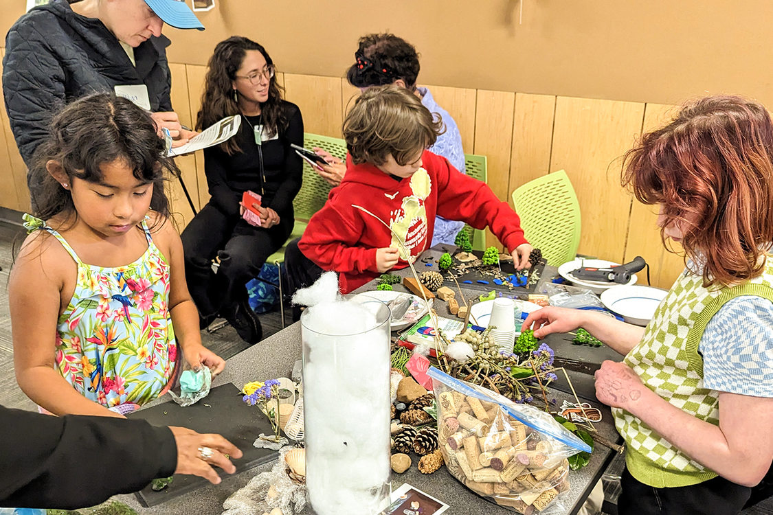 Three children build dioramas of parks out of pinecones, pine need fronds, and other other art supplies at a table. Adults are in conversation behind them.