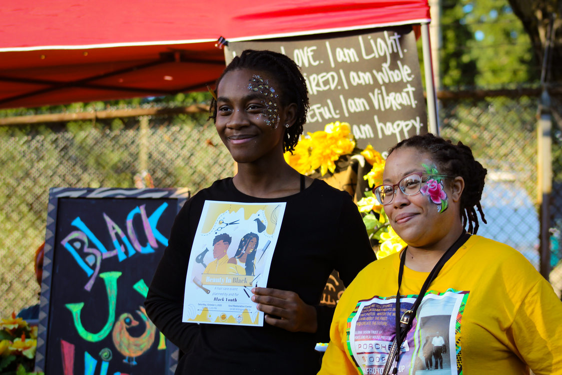Poet La Toya Hampton and youth poet Alyssa Calhoun smile together for a photo in front of a booth. Their faces are painted with flowers and animal print provided by the festival