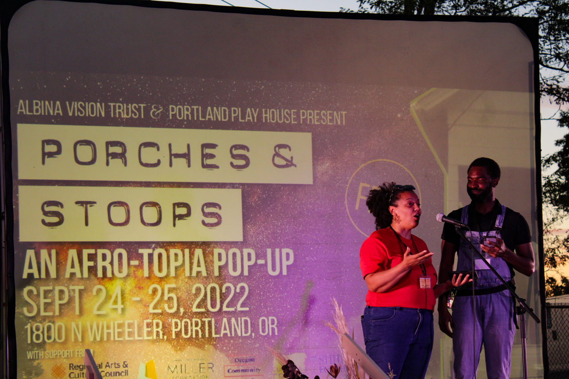 Two presenters speak on stage in front of a large projector screen at the porches and stoops afro-topia pop up. The sun is setting behind them and the screen displays the flyer for the event with a galaxy of stars in the background