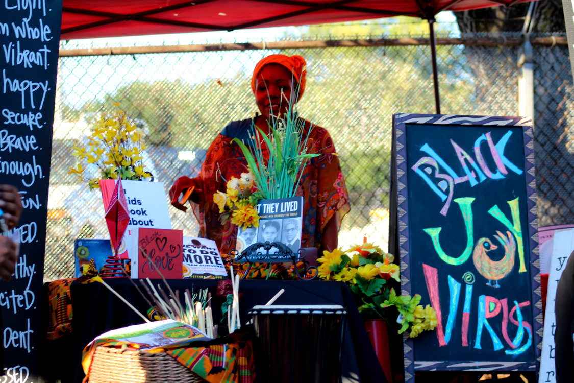 A Black woman stands at her installation at the Afro-topia pop-up event. A colorful sign says "Black Joy Virus" and books, flowers and colorful cloth surround the booth