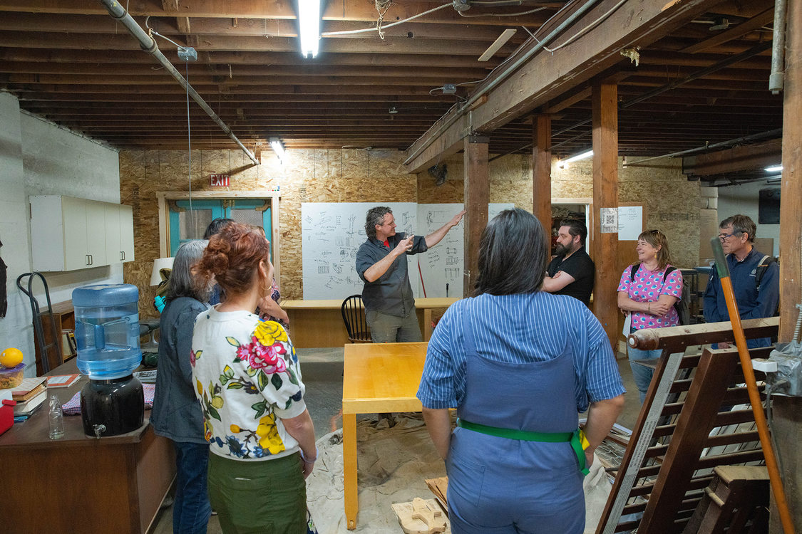 A group of people tour a carpentry studio, a refinished table sits in the middle of the tour group