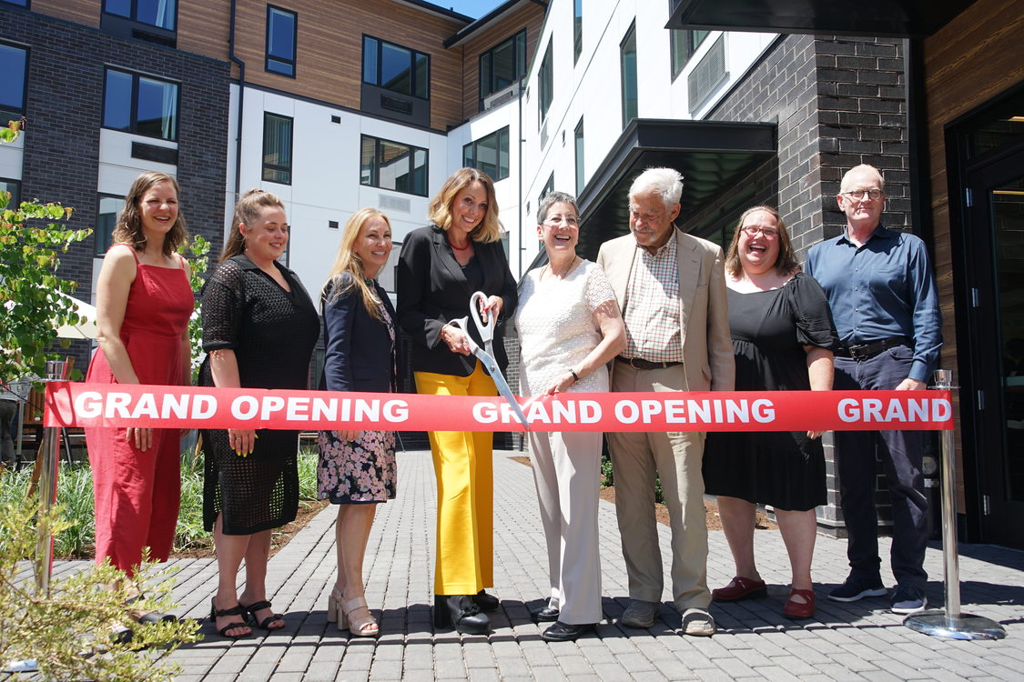 Eight people posing for a photograph jbefore cutting a red "grand opening" ribbon with giant scissors.