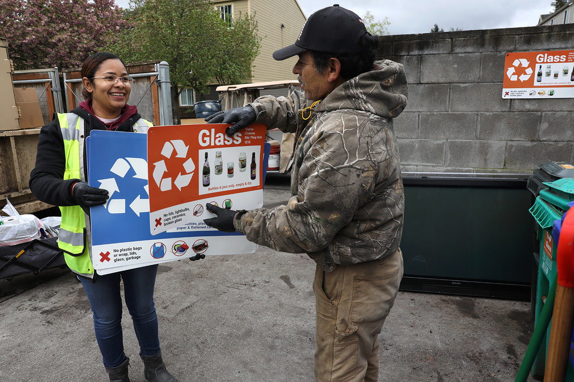 A woman hands large recycling decals to a man in the recycling area at an apartment complex