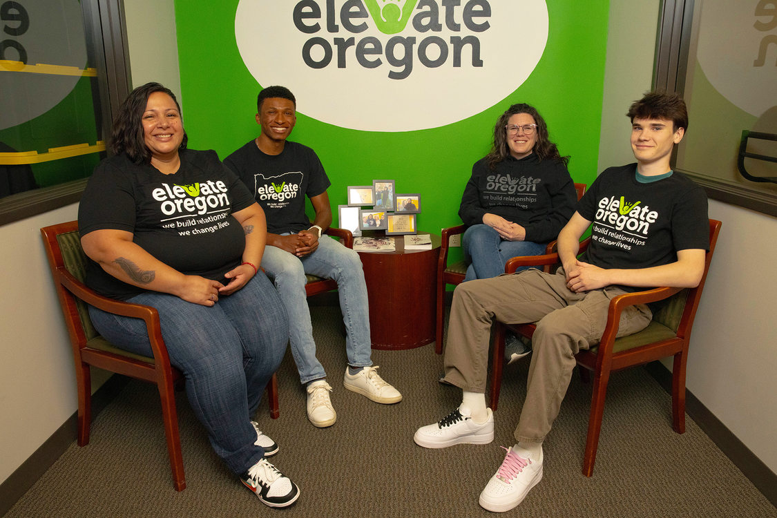 Four people sit in chairs in a hallway at the nonprofit Elevate Oregon. The logo for Elevate Oregon is painted on the wall behind the group.