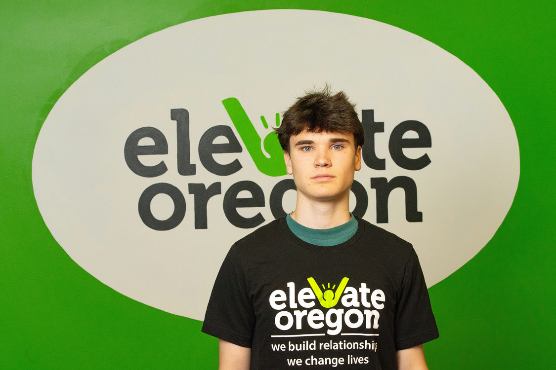 A young person stands in front of wall at Elevate Oregon. The logo for the nonprofit is on the wall behind the person and on their shirt.