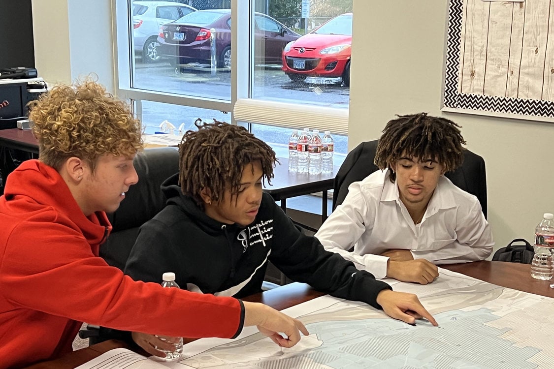 Three young people sit at a conference table looking at a map. Two of them point their fingers at places on the map