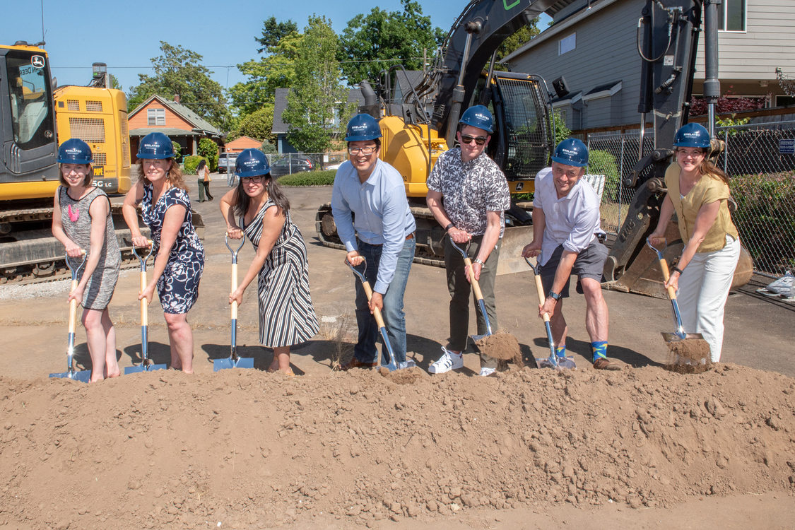 Metro Councilor Duncan Hwang stands alongside six other people wearing hard hats and scooping shovels of dirt with two large construction diggers in the background.