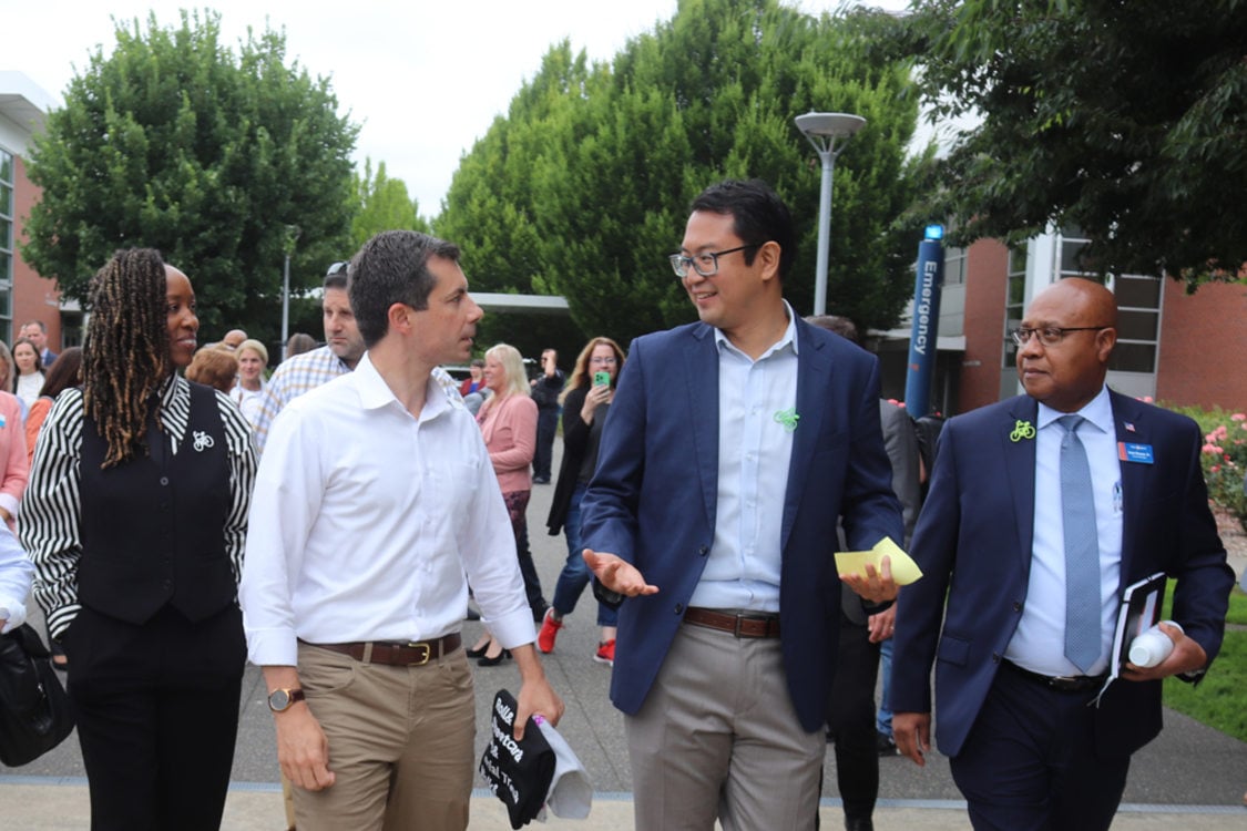 Metro Councilor Duncan Hwang walks alongside Transportation Secretary Pete Buttigieg and other people on a sidewalk at PCC's Southeast campus on 82nd Avenue.