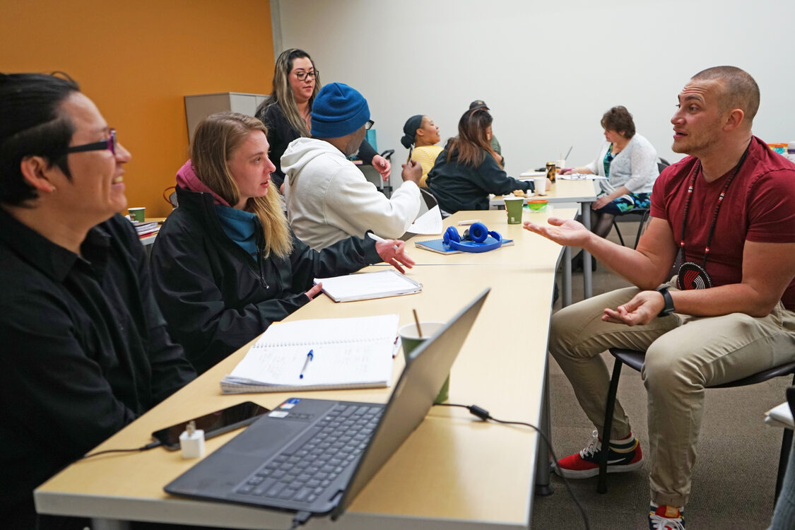 Man in maroon shirt talking to two people at a long table in a classroom setting, with more students in background