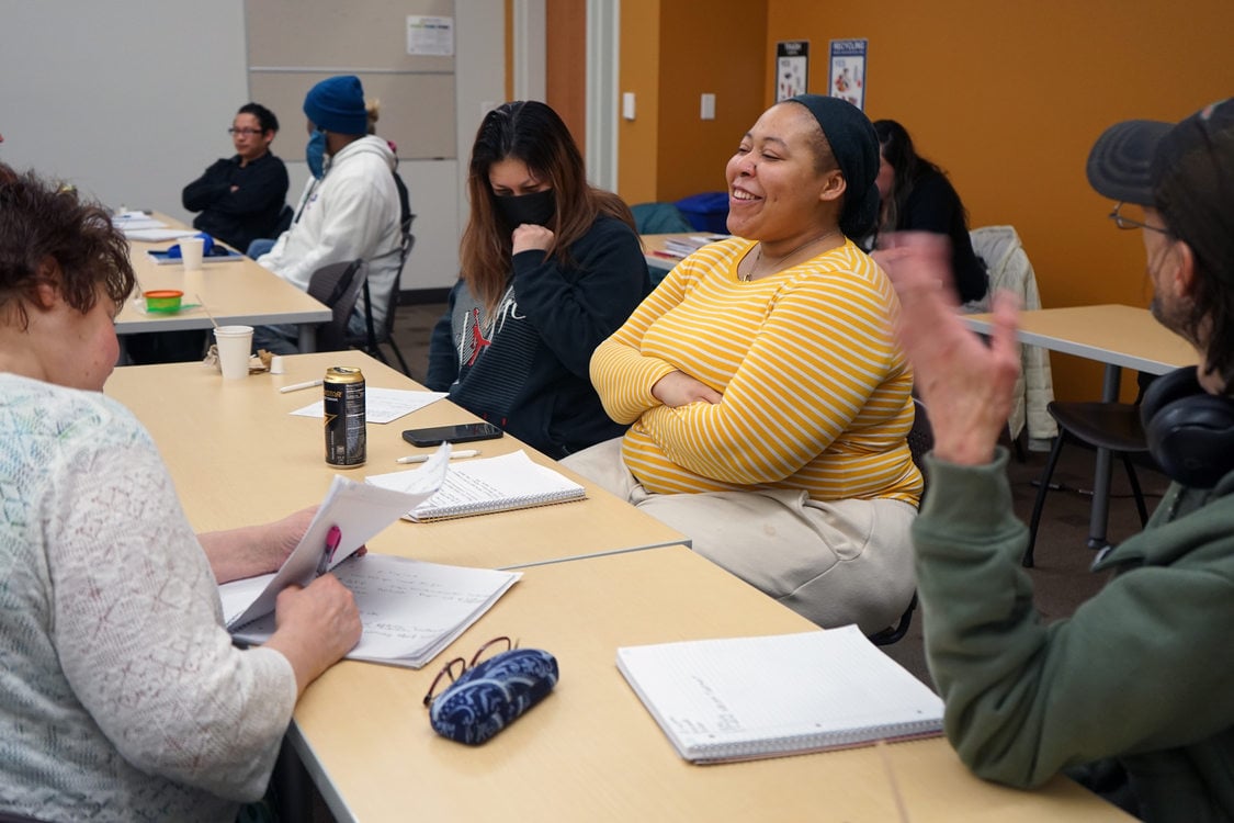 Adult students working at a long table in a classroom setting. A woman in a yellow top is in the center and is smiling with her arms folded.