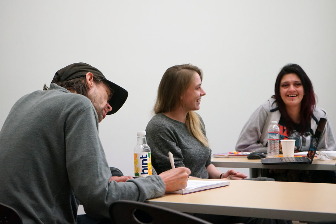 Three adult students in a classroom setting. One is writing in a notebook and the other two are conversing and laughing. 