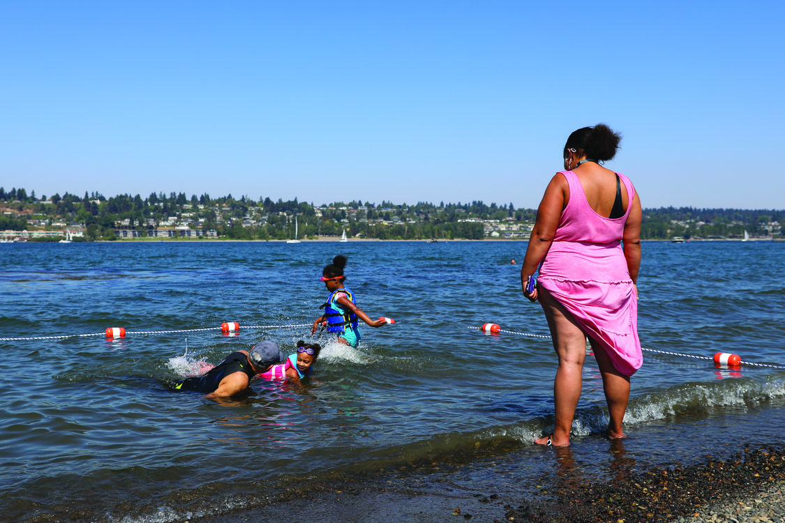 A woman watches from the shore as a swimming instructor teaches two children how to swim in the water. 