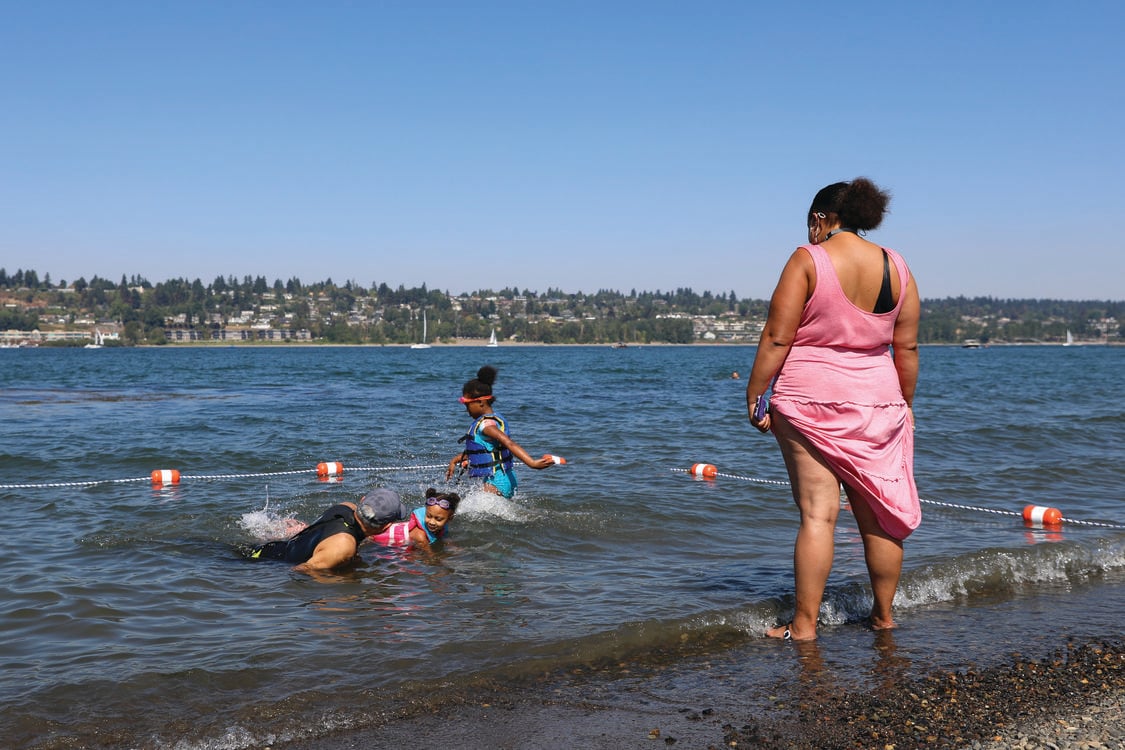 A woman watches from the shore as a swimming instructor teaches two children how to swim in the water. 