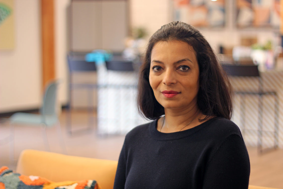 Woman with black hair and shirt looking at the camera, sitting in an indoor space