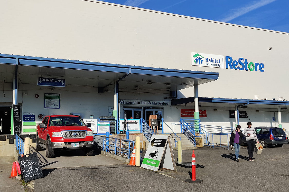 Exterior of a white habitat for humanity ReStore warehouse. A red pickup truck backs into an unloading bay and the sky is bright blue behind the building