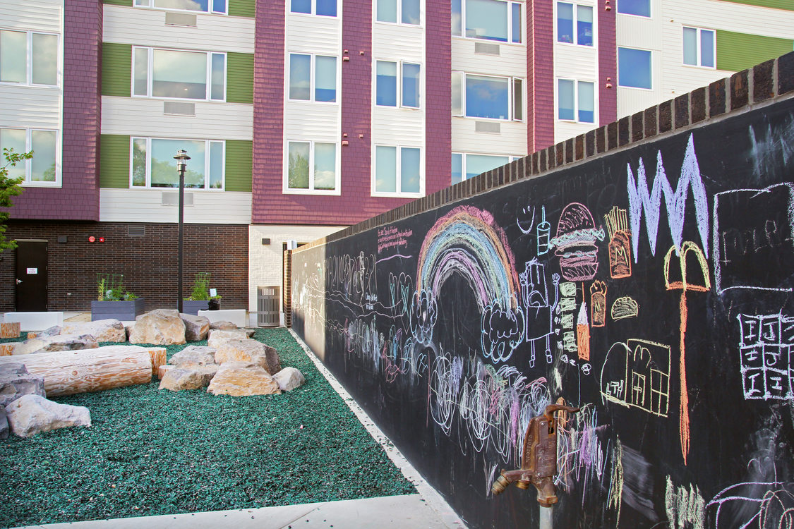 Chalkboard wall with colorful chalk drawkings adjacent to a playground with turf and faux logs, and a multistory apartment building