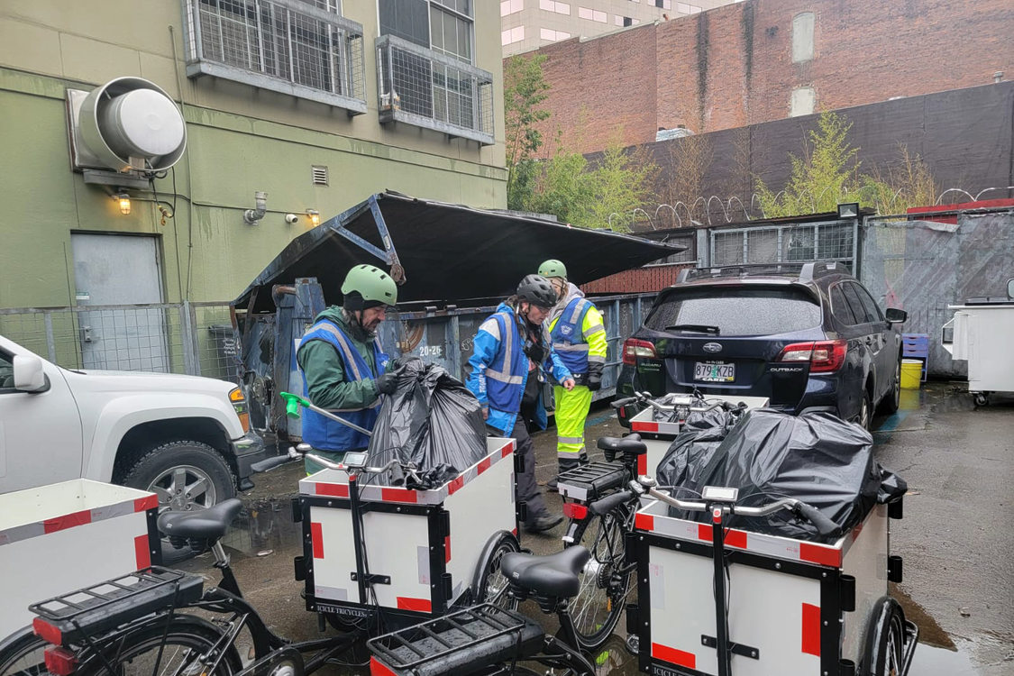 Three people wearing helmets and hi-viz safety gear gather outside next to electric cargo tricycles