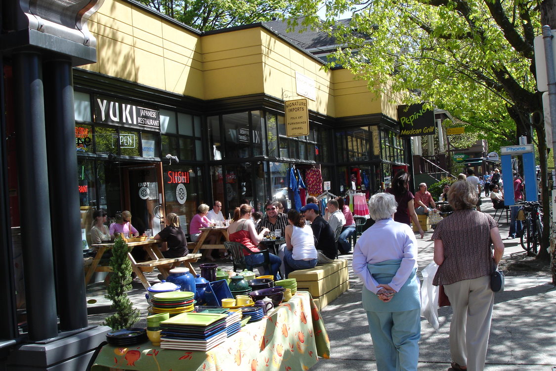 Two people with their backs to the camera stroll through a busy commercial district. To their left, a large group of people sit and the outdoor tables of a dining establishment.